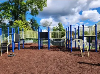 A vibrant, new playground with blue posts and yellow railings on fresh brown wood chips, under a sunny sky.
