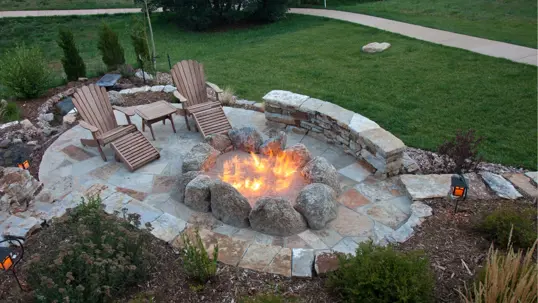 An inviting outdoor fire pit area at dusk. A stone patio features a roaring fire pit, two wooden chairs, and landscaped surroundings.