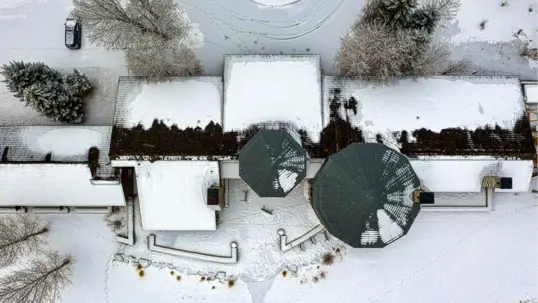 An aerial view shows a snow-covered property with a house, two large dark circular structures, and trees.