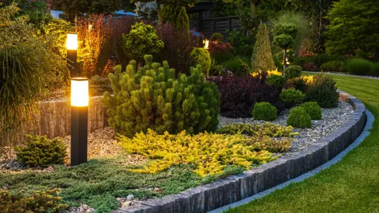 A stunning garden bed at night, beautifully lit with modern landscape lights highlighting diverse plants and shrubs.