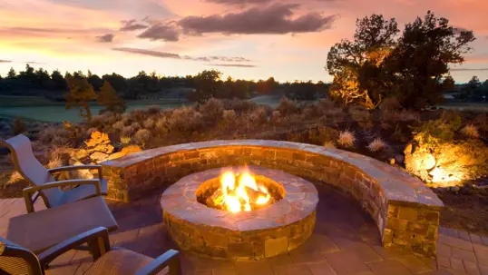 A beautiful stone fire pit with a curved seating wall on a patio at sunset, ideal for igniting outdoor experiences. Landscaped grounds and a distant golf course complete the scene.