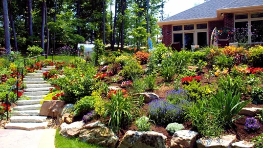 A lush, terraced garden displays a stone staircase leading up to a house. Colorful flowers, plants, and decorative rocks adorn the vibrant landscape.
