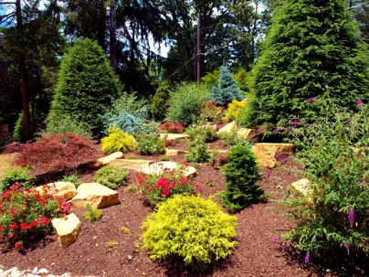 A vibrant garden landscape showcases diverse trees, colorful shrubs with red and yellow flowers, and natural stone accents over mulched ground.