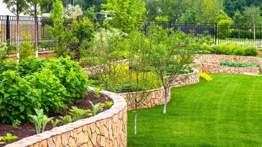 A beautifully landscaped terraced garden features curving stone retaining walls, lush green plants, small trees, and a vibrant manicured lawn.