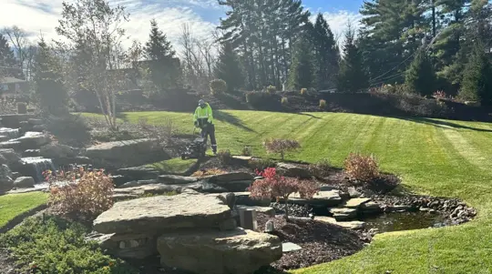 A professional landscaper mows a lush, striped lawn in a beautifully designed backyard featuring a rock waterfall and pond.