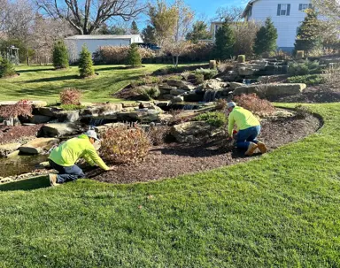 Two landscapers in bright yellow shirts tend to plants beside a beautiful multi-tiered rock waterfall and stream, showcasing a well-maintained water feature.