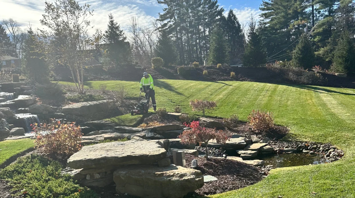 A person in a bright yellow jacket professionally mows a striped green lawn next to a beautiful rock garden with a waterfall feature, showcasing detailed landscaping.