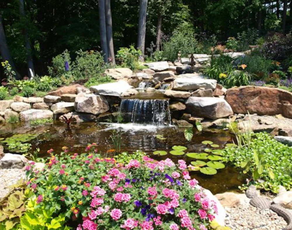 A beautiful multi-tiered rock waterfall flows into a serene pond with lily pads and vibrant foreground flowers.